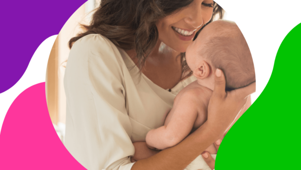 Frau mit braunen Haaren hält ihr Baby lächelnd in der Hand und genießt die Zeit nach der Geburt