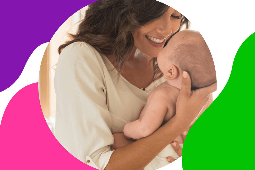 Frau mit braunen Haaren hält ihr Baby lächelnd in der Hand und genießt die Zeit nach der Geburt